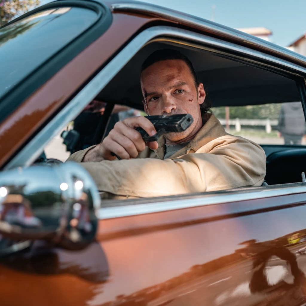Alban Lenoir as Gab in Les orphelins produced by Inoxy films A man in a beige jacket, shooting a pistol through the open window of a red car - Alban Lenoir as Gab in Les orphelins produced by Inoxy films